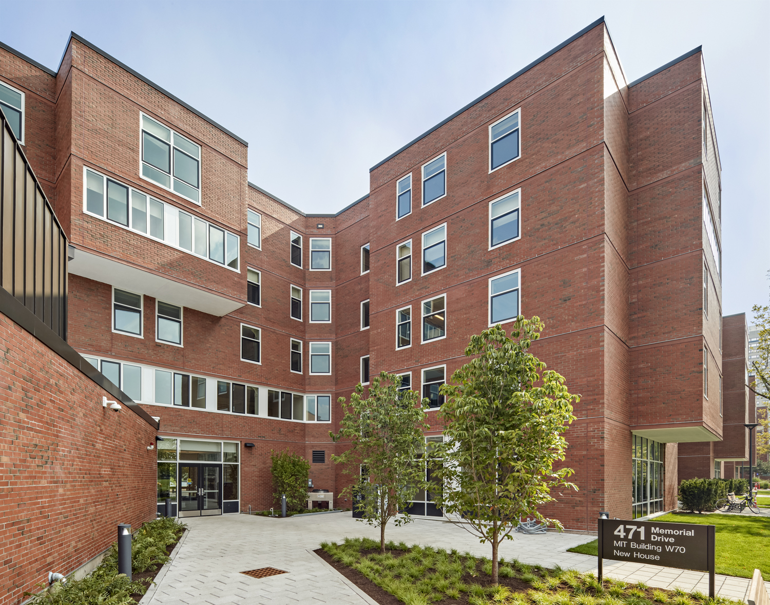 Front entrance of brick building with windows along the front, greenery in the foreground with New House sign at the bottom right corner, blue skies in the background.