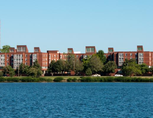 Skyline of brick building, greenery in front of the building, Charles River in the foreground, blue skies in the background.